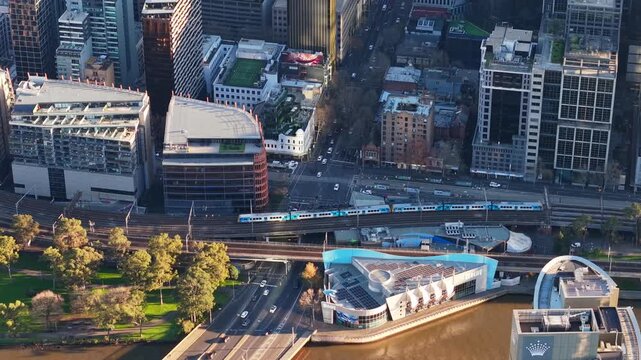 Aerial drone video over Spencer Street in Melbourne CBD, showing trains passing along elevated tracks above road traffic, with parkland, city buildings and the Yarra River