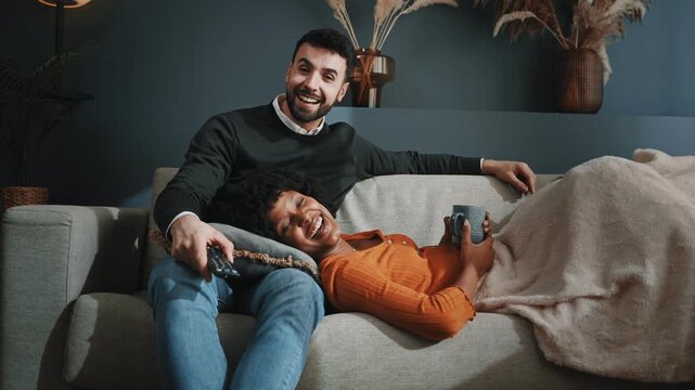 Relaxed multi ethnic couple sitting on sofa watching television show on online streaming video on demand together at home