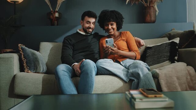 Young couple sitting on sofa using mobile phone app to share social media content at home