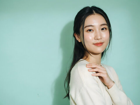 Portrait of a young east asian woman with long dark hair and natural makeup posing against a soft pastel green background, in a studio