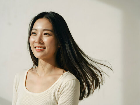 Young asian woman with long dark hair blowing in the wind, smiling, wearing a beige shirt against a plain white wall with soft sunlight shadows