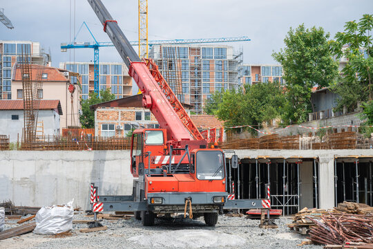 a heavy red mobile crane with its outriggers deployed on an active urban construction site, positioned in front of a concrete foundation and surrounded by building materials.