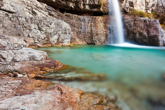 Waterfall cascading into a turquoise pool with autumn leaves on rocky foreground, tourism destination for outdoor nature adventure in the Julian Alps Natural Park, Resia, Friuli Venezia Giulia, Italy.