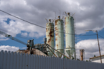 three tall, rusting industrial storage silos and an aging crane stand behind a white corrugated fence under a dramatic, cloud-filled sky. © Andrey
