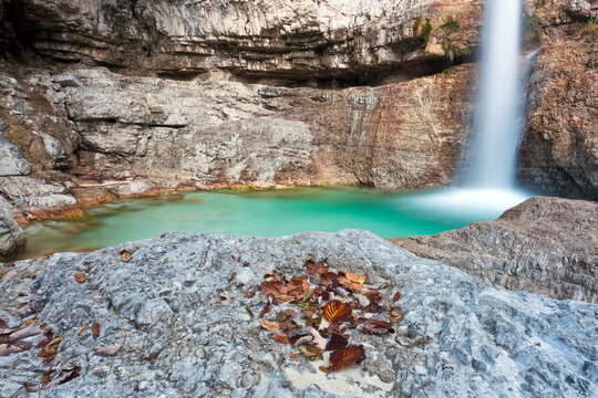 Waterfall cascading into a turquoise pool with autumn leaves on rocky foreground, tourism destination for outdoor nature adventure in the Julian Alps Natural Park, Resia, Friuli Venezia Giulia, Italy.