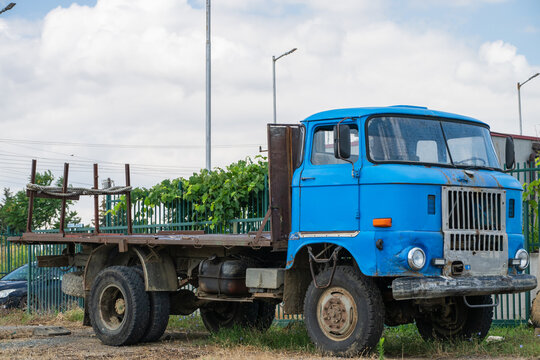 a vintage blue cab-over flatbed truck is parked on a dirt and grass lot.