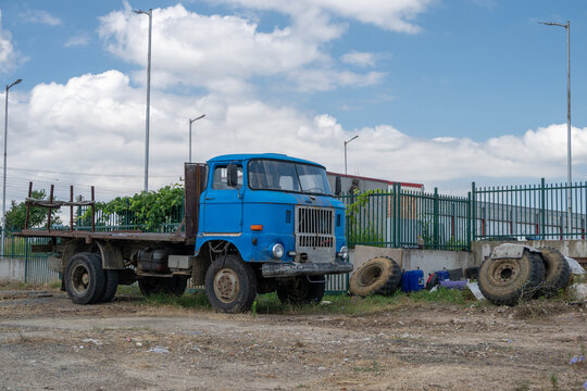 a vintage blue cab-over flatbed truck is parked on a dusty dirt lot next to several large, discarded heavy-duty tires