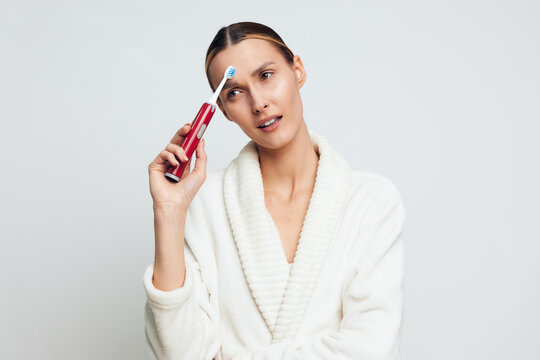 A young woman in a white bathrobe holds an electric toothbrush near her forehead with a confused expression. The plain background emphasizes personal hygiene and morning routine concepts.