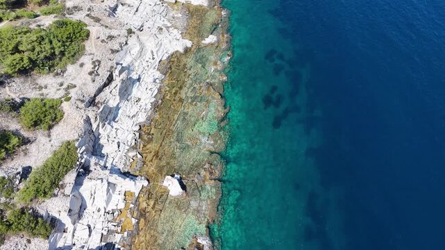 Vertical aerial view drifting along a rugged white stone shoreline bordered by turquoise shallows and deep blue sea.