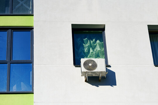 Outdoor air conditioner compressor unit mounted on white wall below window with green curtains. AC split system outdoor unit installed on white plastered facade. Air conditioner unit on building wall