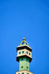 Obraz premium Mosque Minaret with Dome Low-angle view of a modern mosque minaret tower featuring a golden dome and green windows against a vibrant clear blue sky background for religious design.