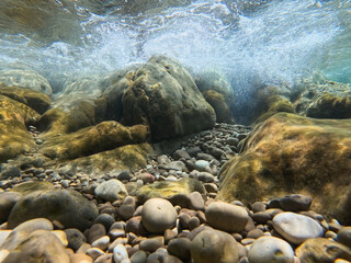 Fototapeta premium Underwater view of clear turquoise sea water with small pebbles, large rocks, and breaking waves along the coastline of Saint-Jean-Cap-Ferrat, France