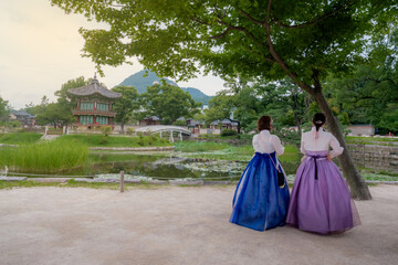 Fototapeta premium Rear view of two young women wearing traditional Korean Hanbok dresses standing under a tree, looking at the beautiful pond and Hyangwonjeong Pavilion at Gyeongbokgung Palace in Seoul, South Korea.