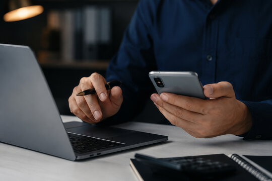 Businessman working late using laptop holding smartphone pen calculator desk. Multitasking finance accounting overtime remote online business communication mobile technology concept. 