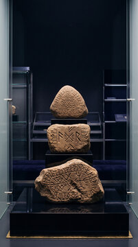 Vertical shot of ancient Norse runestones stacked in a museum display case. Weathered stones with carved runic inscriptions and Celtic knotwork symbols. Archaeological artifact exhibition