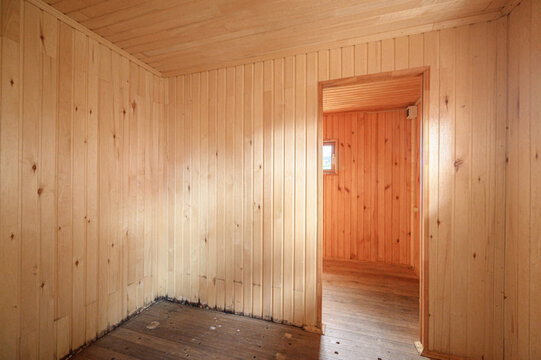 wooden interior features vertical plank cladding on walls and ceiling, a dark wooden floor, and an open doorway leading to a room with a small window