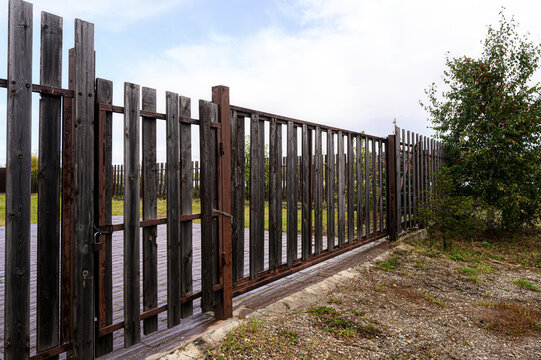 sturdy fence and sliding gate combine weathered wooden slats with rusted metal framing. sparse grass and a leafy tree border a gravel edge under a pale sky