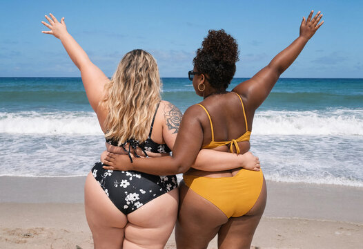 Two Diverse Plus Size Female Friends in Bikinis Embracing on a Sunny Beach looking at Ocean