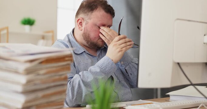 Man accountant stressed at computer with paperwork. He rubs eyes by monitor, feeling tired while tracking budget deadlines as a busy office worker. Clear concept of work burnout and stress.