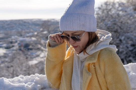Young woman wearing yellow coat and white beanie adjusting sunglasses while standing outdoors in snowy winter landscape