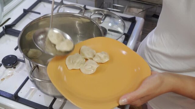 A hand holds a yellow plate while scooping fresh dumplings from a boiling pot on the stove. Serving this hot meal brings a satisfying, comforting feeling of happy anticipation.