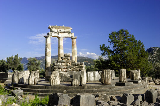 The Tholos of Athena Pronaia at Delphi, Greece, stands under a clear blue sky, showcasing its ancient Doric columns and weathered stone ruins amidst lush greenery and distant mountains.