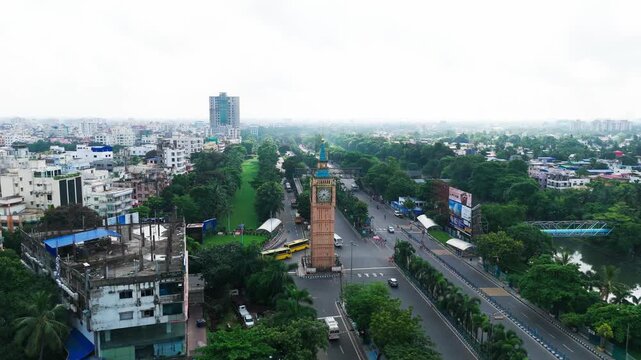 Daytime aerial drone video of Big Ben Clock Tower replica in Lake Town Kolkata, surrounded by green trees, road traffic intersection, and city landscape in West Bengal.