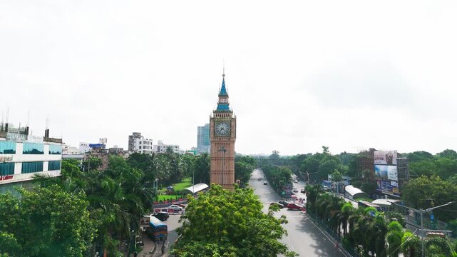 Daytime aerial drone video of Big Ben Clock Tower replica in Lake Town Kolkata, surrounded by green trees, road traffic intersection, and city landscape in West Bengal.
