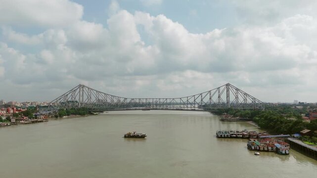 Daytime aerial view of the iconic Howrah Bridge spanning across the Hooghly River in Kolkata under a cloudy sky, showcasing cantilever steel architecture and river landscape.