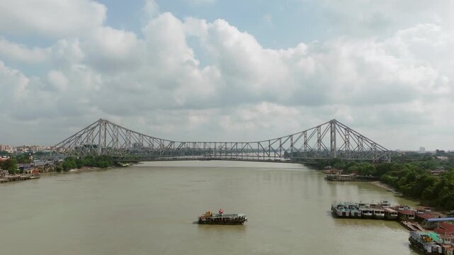 Daytime aerial view of the iconic Howrah Bridge spanning across the Hooghly River in Kolkata under a cloudy sky, showcasing cantilever steel architecture and river landscape.