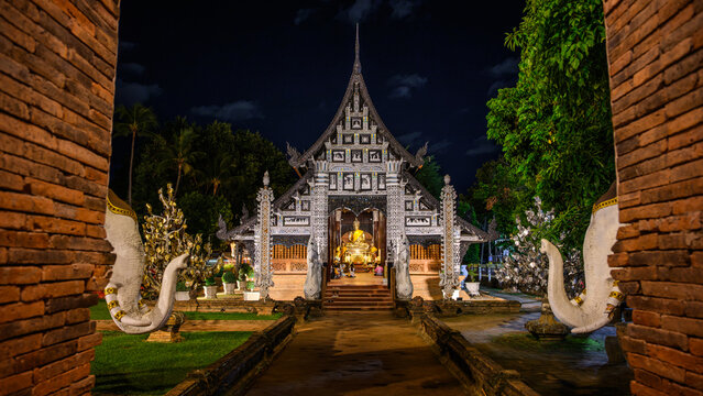 Ancient Lanna style teak wooden viharn of Wat Lok Moli illuminated at night, framed by brick archways in Chiang Mai, Thailand. Historic Buddhist temple landmark.