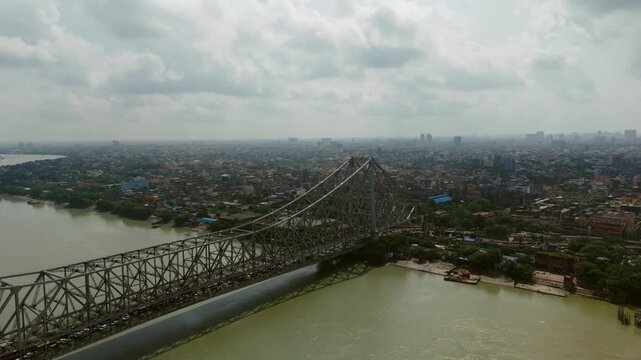 Daytime aerial view of the iconic Howrah Bridge spanning across the Hooghly River in Kolkata under a cloudy sky, showcasing cantilever steel architecture and river landscape.