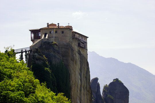 The Monastery of Great Meteoron perched atop a rock formation in Meteora, Greece, with lush green trees in the foreground and hazy mountains in the background.