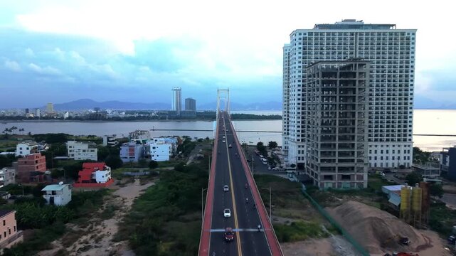 Drone flying above a modern bridge toward the Da Nang skyline with dramatic morning clouds over the Han River.