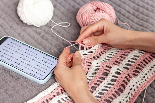 Close up of female hands crocheting pink and white cotton fabric with help of digital pattern chart on mobile phone screen.