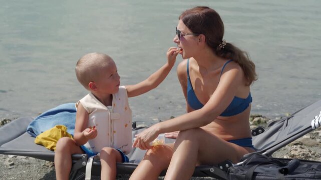 A loving mother feeds sweet treats to her happy son in a life vest. They sit on a beach lounger by a calm lake with green algae.