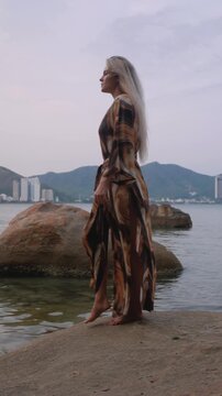 Fashion model wearing abstract print beach kimono posing on rocky tropical shoreline by calm ocean.