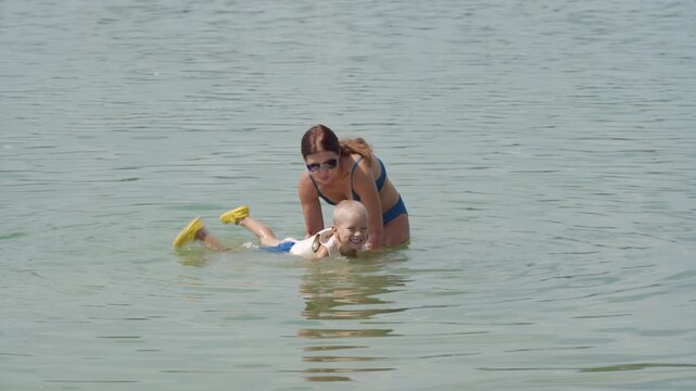 A loving mother gently supports her happy son as he swims. They share a joyful moment in a calm, warm lake filled with green algae.