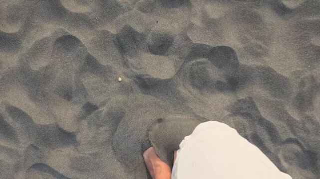 High angle view of bare feet walking on soft grey beach sand during a sunny day.