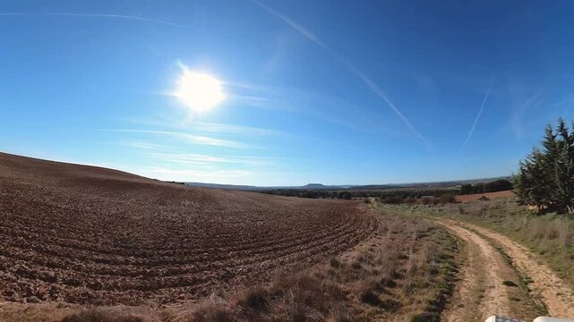Classic Land Rover Defender drives along a narrow dirt track between cultivated fields in a wide open valley, rural landscape in the province of Soria, Spain. Forward and side view of the road during 