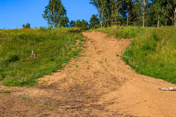 Obraz premium Sandy dirt path winding up a lush green grassy hillside under a clear blue sky, showing traces of environmental impact and rough terrain on a sunny summer day
