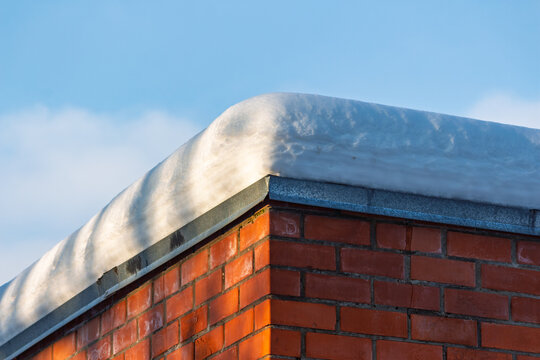 Thick layer of fresh white snow rests heavily on the sharp corner of a weathered red brick building roof against a crisp clear blue winter sky during a sunny day