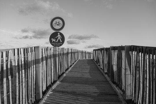 Black and white wooden boardwalk path leading through coastal dunes toward Labenne-Oc&eacute;an in the Landes, France, with regulatory signs. Monochrome landscape.
