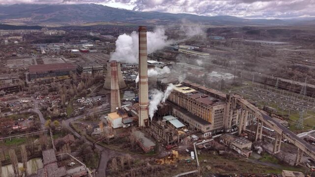 Cinematic aerial view of a thermal power station with massive cooling towers, smokestacks and industrial infrastructure. Steam rises into a dramatic cloudy sky while the nearby city highlights the rel