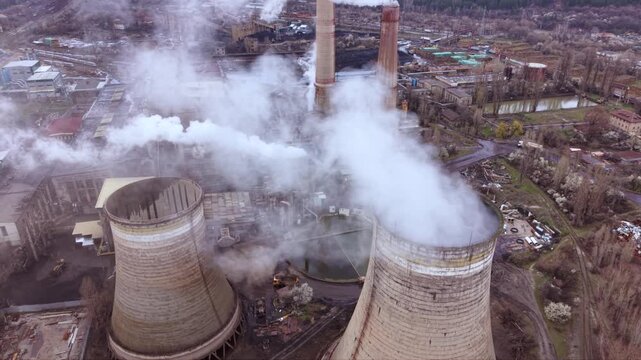 Cinematic aerial view of a thermal power station with massive cooling towers, smokestacks and industrial infrastructure. Steam rises into a dramatic cloudy sky while the nearby city highlights the rel