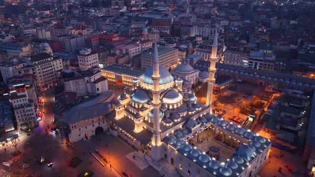 Cinematic aerial view of the New Mosque (Yeni Cami) illuminated during blue hour overlooking Emin&ouml;n&uuml; Square in the heart of Istanbul, Turkey. Historic Ottoman architecture, lively streets and the vibr