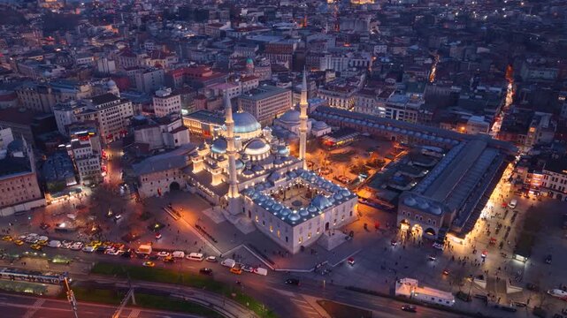Cinematic aerial view of the New Mosque (Yeni Cami) illuminated during blue hour overlooking Emin&ouml;n&uuml; Square in the heart of Istanbul, Turkey. Historic Ottoman architecture, lively streets and the vibr