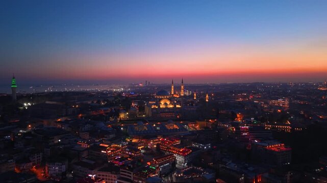 Cinematic aerial view of the illuminated S&uuml;leymaniye Mosque during sunset in Istanbul, Turkey. The iconic Ottoman landmark dominates the historic skyline while city lights emerge beneath a colorful tw