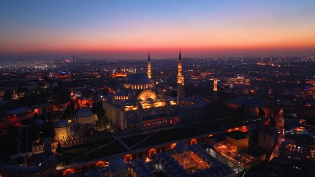 Cinematic aerial view of the illuminated S&uuml;leymaniye Mosque during sunset in Istanbul, Turkey. The iconic Ottoman landmark dominates the historic skyline while city lights emerge beneath a colorful tw