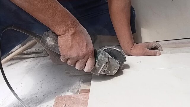 A man's hands cutting a ceramic floor tile with an angle grinder during indoor renovation. Construction worker trimming tiles with precision for flooring installation.